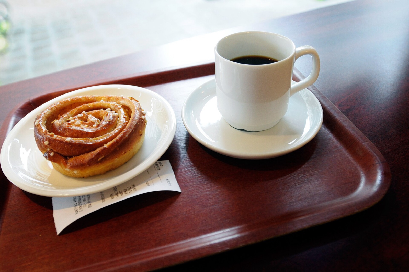 Un kanelbulle sur une assiette et une tasse de cafe noir sur un plateau, dans un cafe suedois