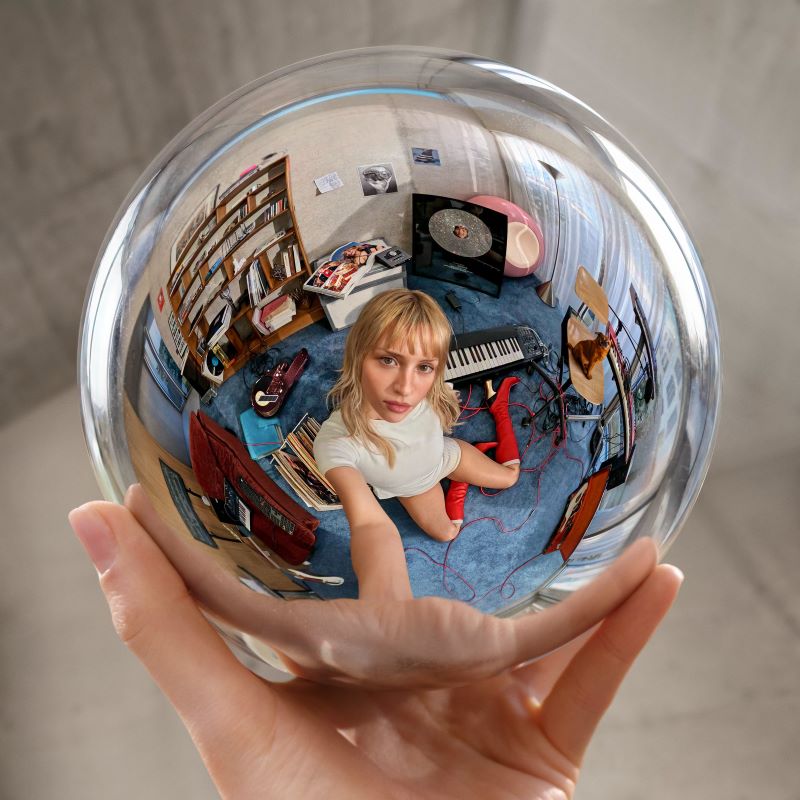 Pochette de l'album Instinct d'Angèle : la chanteuse assise dans une pièce reflétée dans une sphère de verre tenue par une main