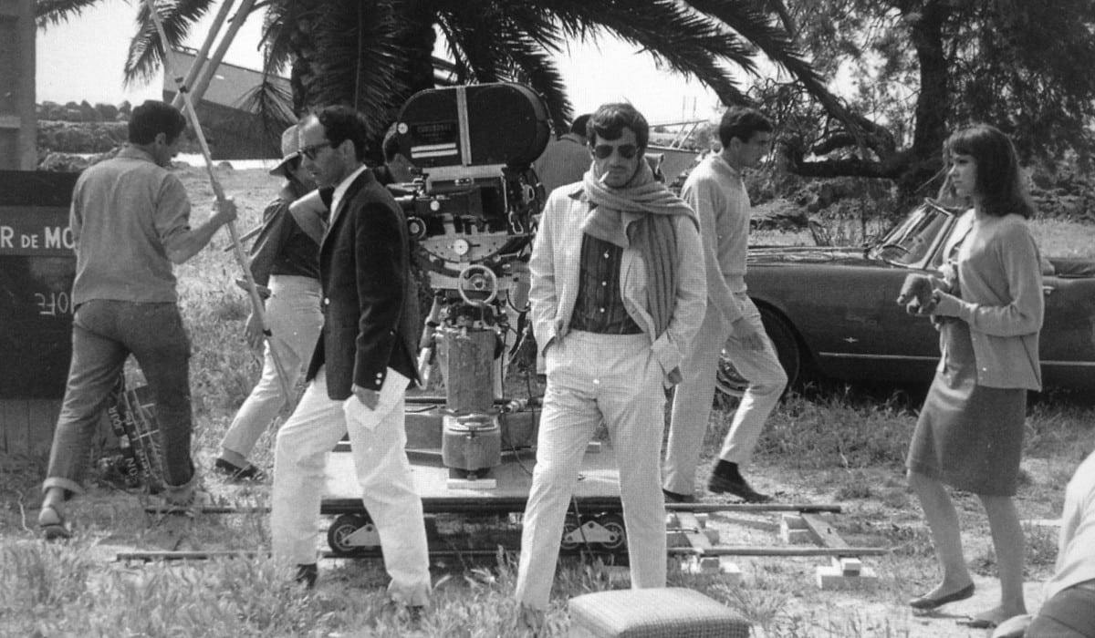 Jean-Paul Belmondo sur un tournage, lunettes de soleil et écharpe, devant une caméra