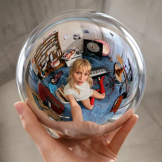 Pochette de l'album Instinct d'Angèle : la chanteuse assise dans une pièce reflétée dans une sphère de verre tenue par une main