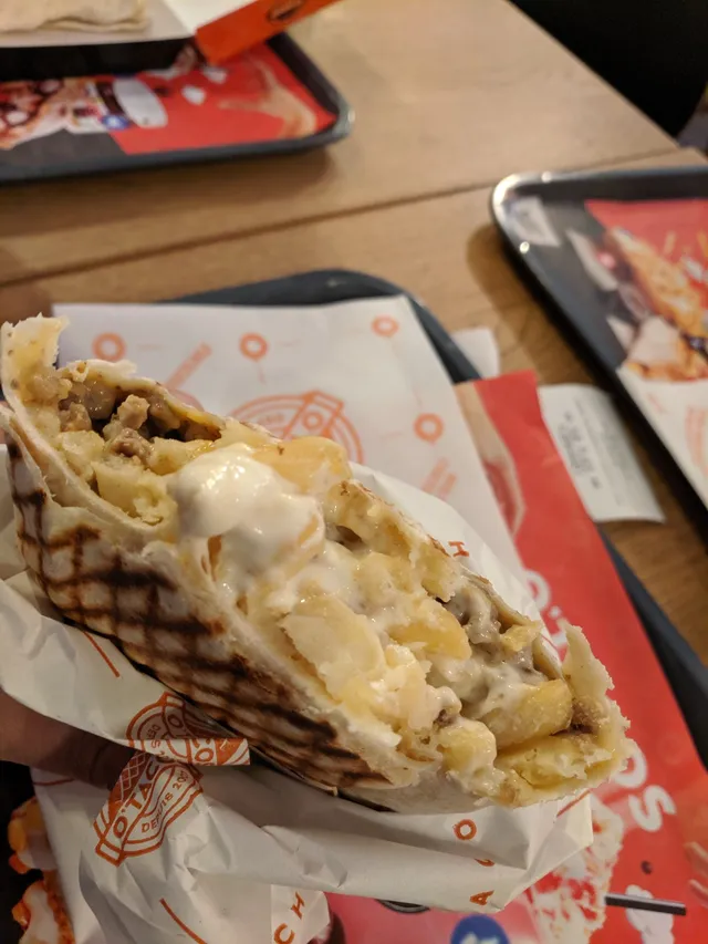 Un french tacos gratiné ouvert, galette de blé grillée garnie de viande, de frites et de sauce fromagère fondante, dans un emballage O'Tacos
