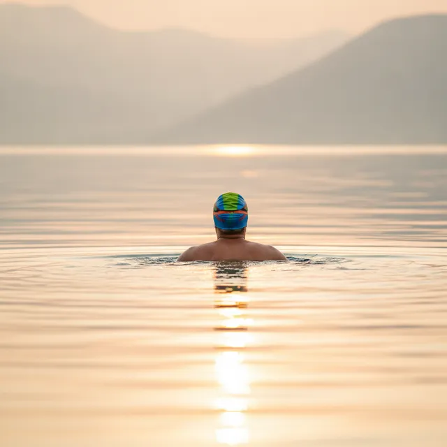 Un nageur de dos en eau libre dans un lac calme au lever du soleil, bonnet coloré, reflets dorés sur l'eau
